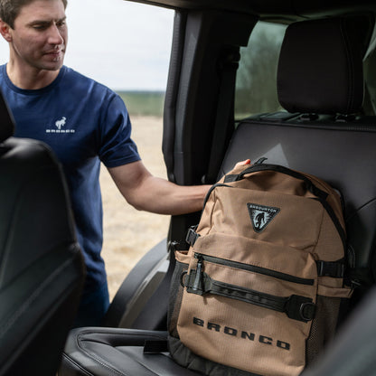Man in a blue shirt with a 'Bronco' logo placing a brown backpack on the back seat of a vehicle.