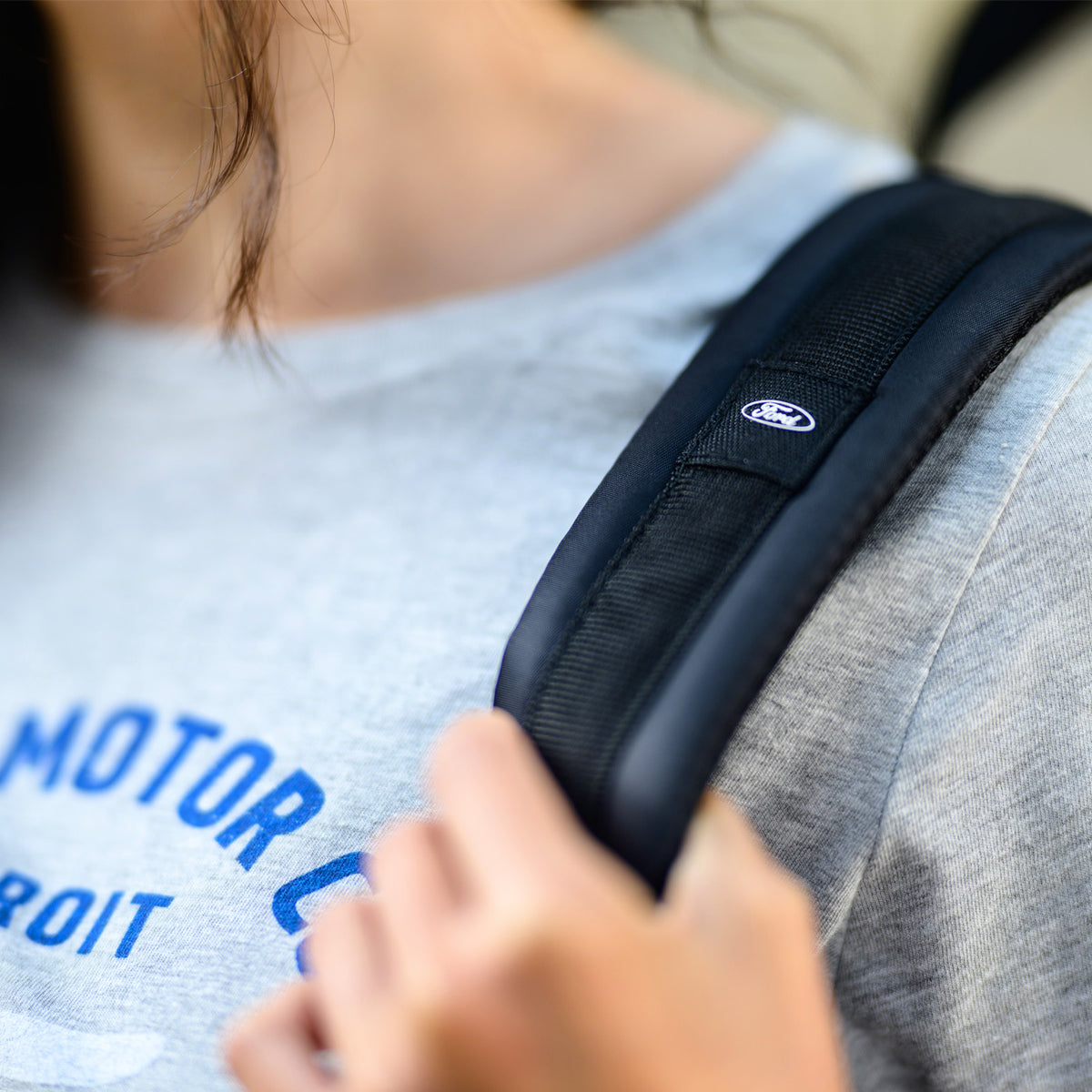 Person wearing a black backpack strap with a visible Ford logo, wearing a gray shirt with blue text.