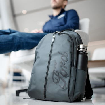 Gray backpack with 'Ford' logo on a surface, person sitting in the background