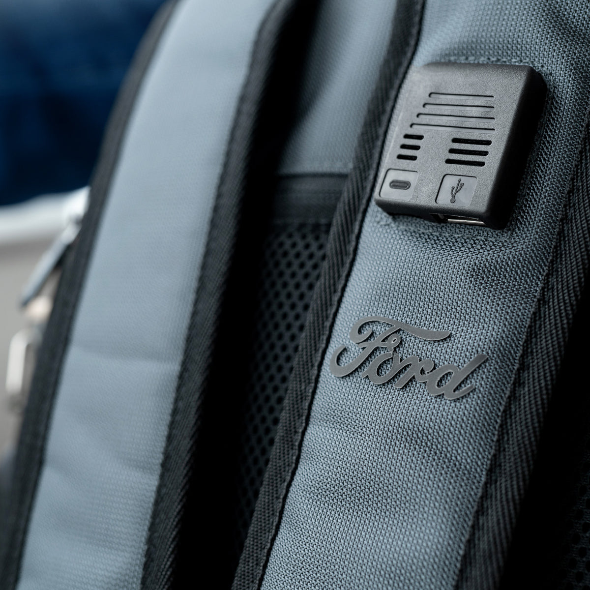 Gray backpack with Ford logo and USB port on a blurred outdoor background