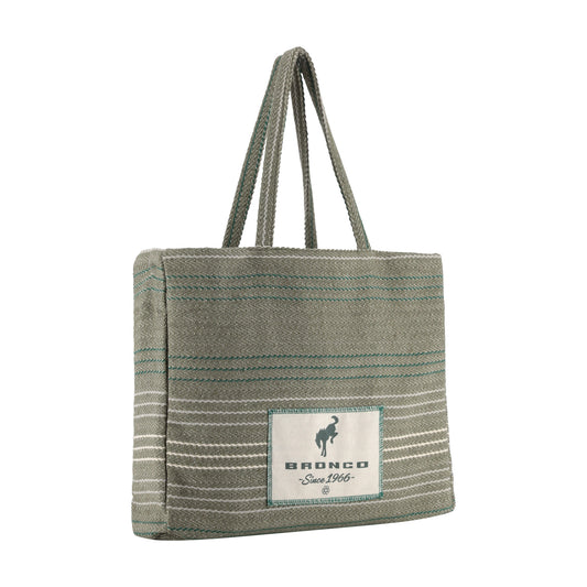 A woven tote bag in a muted sage green color with thin teal and white horizontal stripes, featuring a white rectangular patch on the front with the brand name 'BRONCO' and a horse logo.