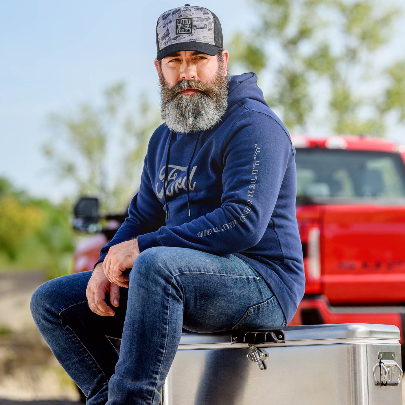 Man with a beard wearing a blue hoodie and cap sitting on a toolbox with a red truck in the background.