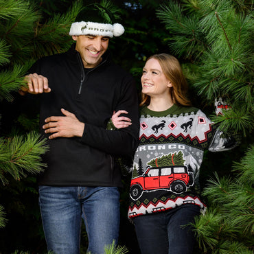 a man and a women holding arms walking out of pine tree in a bronco holiday sweater and a bronco santa hat