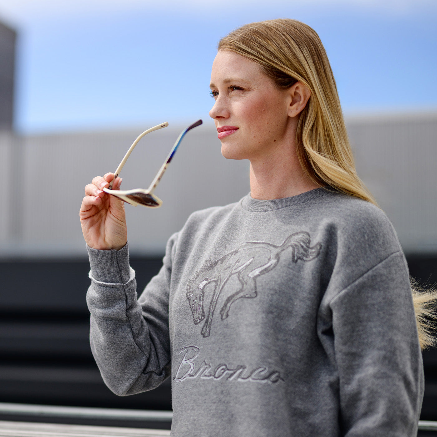 Woman wearing a gray sweatshirt with a horse design, holding sunglasses.