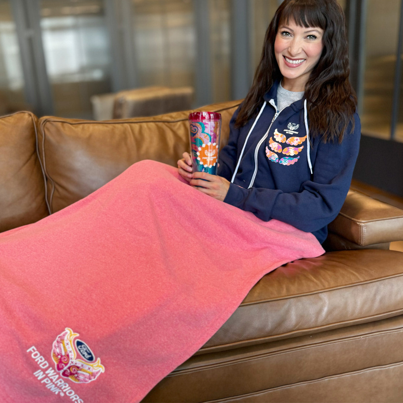 women with brown hair in navy blue sweatshirt holder tumbler wrapped in pink Ford warriors in pink blanket sitting on brown couch