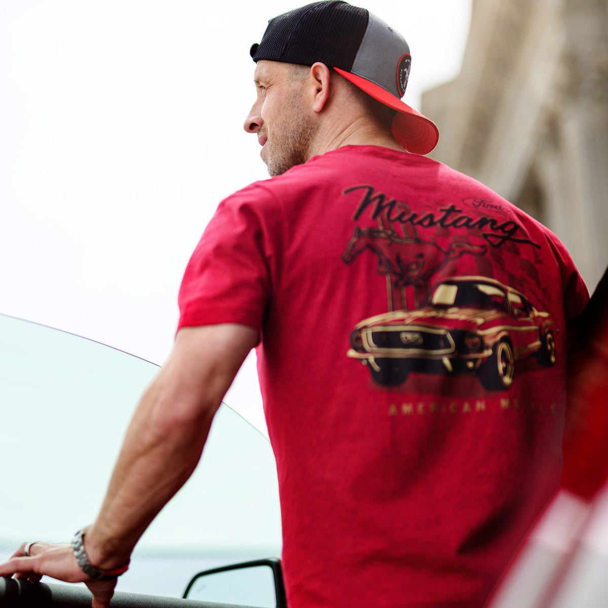 Man wearing a red t-shirt with a car graphic and 'Mustang' text, standing next to a car.