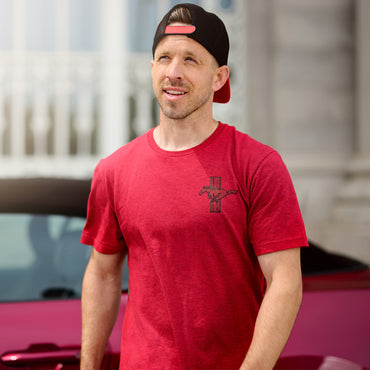 Man wearing a red t-shirt with a logo and a black cap, standing in front of a pink car.