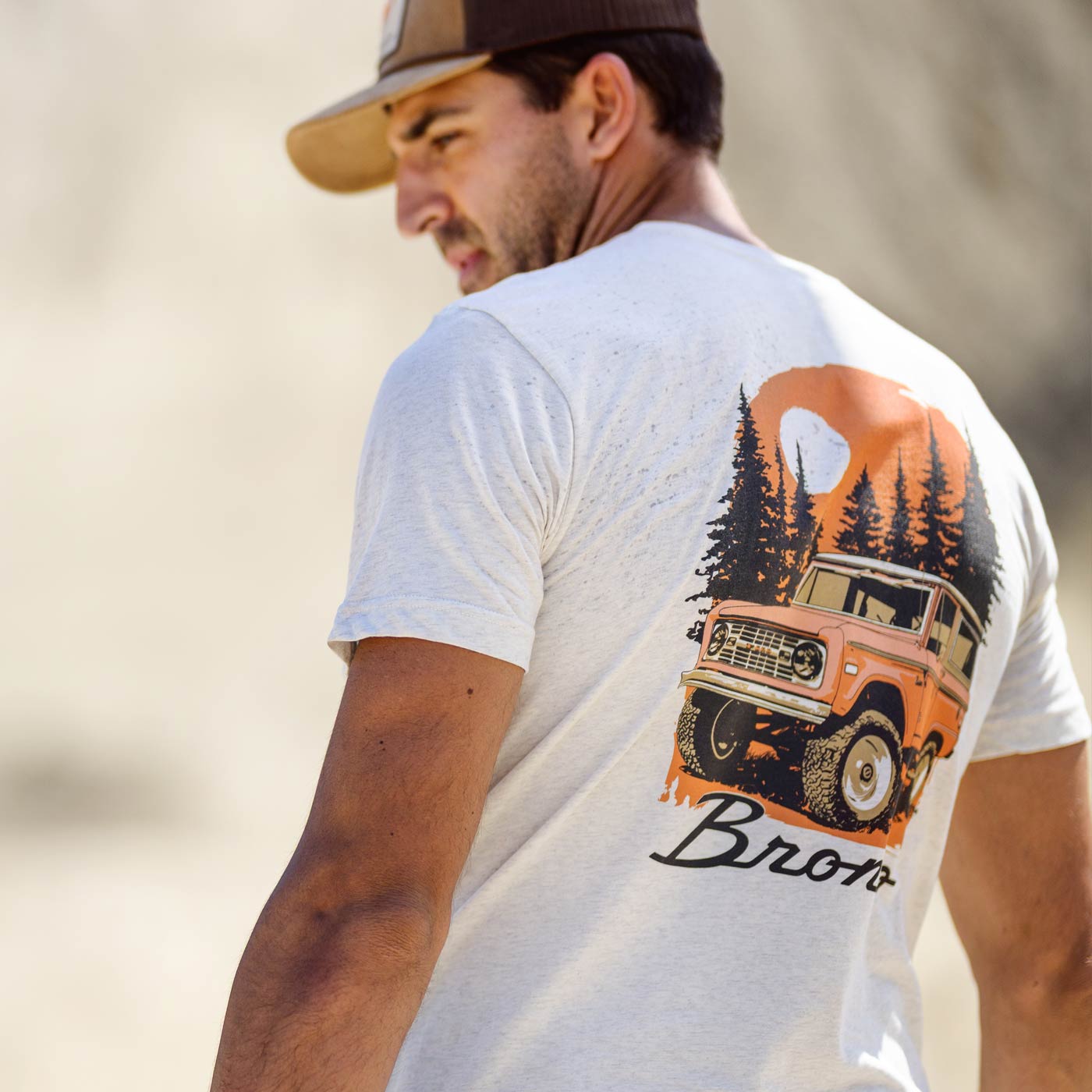 Man wearing a white t-shirt with a graphic of a vintage truck and trees on the back, set against a blurred natural background.