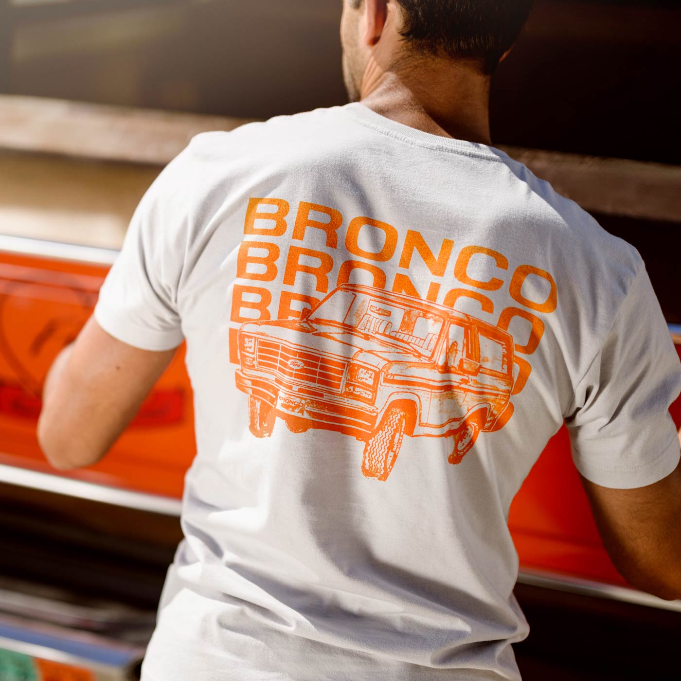 Person wearing a white t-shirt with orange 'Bronco' text and graphic, standing next to a red vehicle.