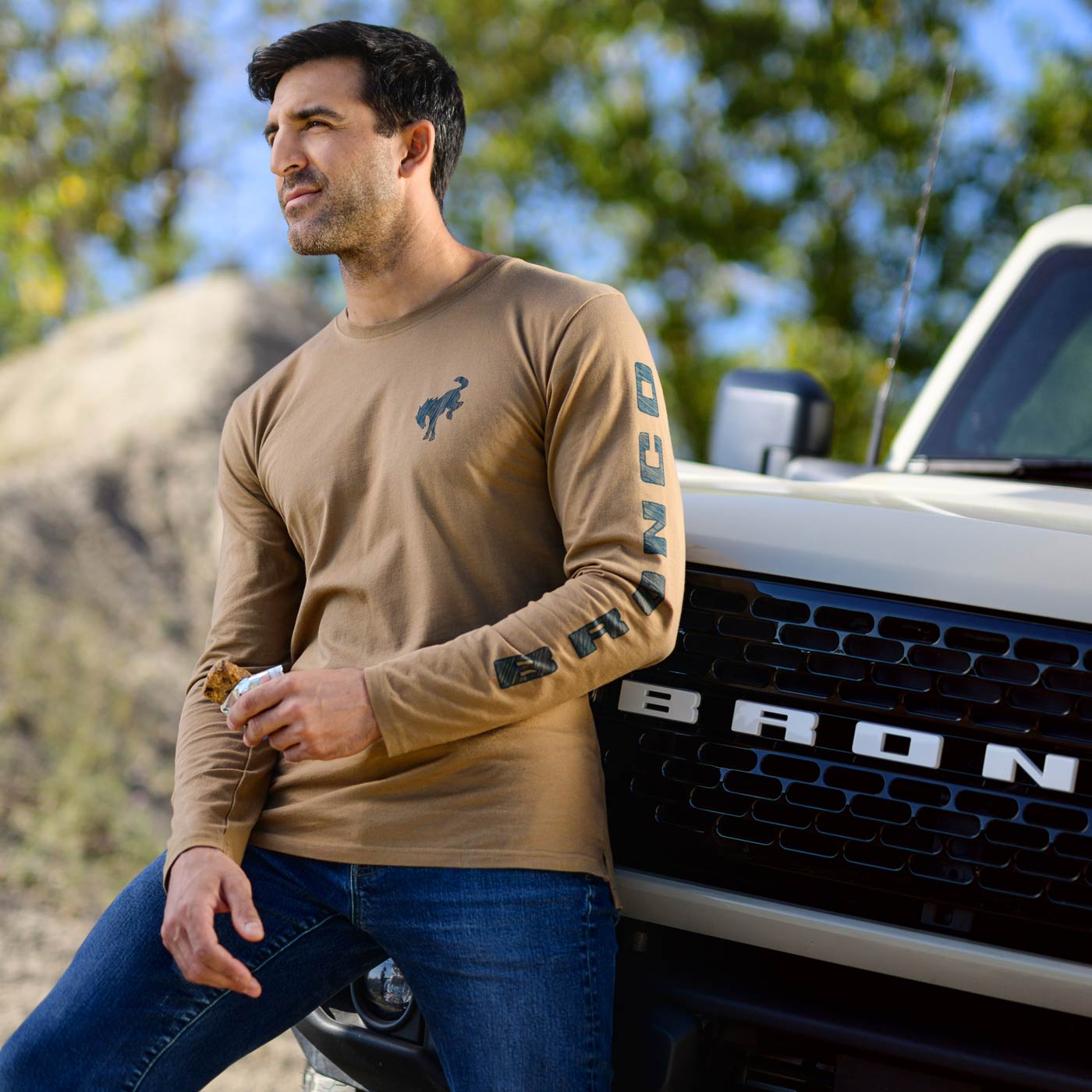 Man wearing a tan long-sleeve shirt with a logo, sitting next to a white truck with 'Bronco' branding.