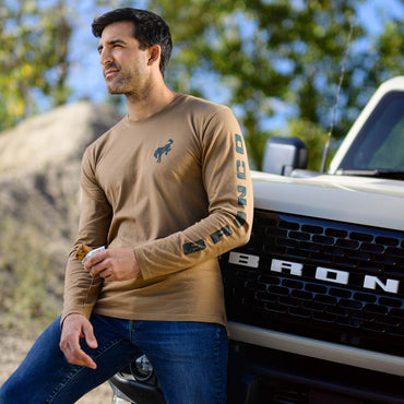 Man wearing a tan long-sleeve shirt with a logo, sitting next to a white truck with 'Bronco' branding.