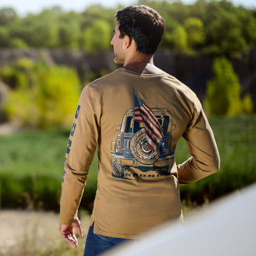 Man wearing a tan long-sleeve shirt with a graphic design of a truck and American flag, standing outdoors.