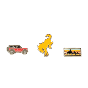 Three enamel pins on a white background: a red car, a yellow dinosaur, and a rectangular badge with mountains and text.