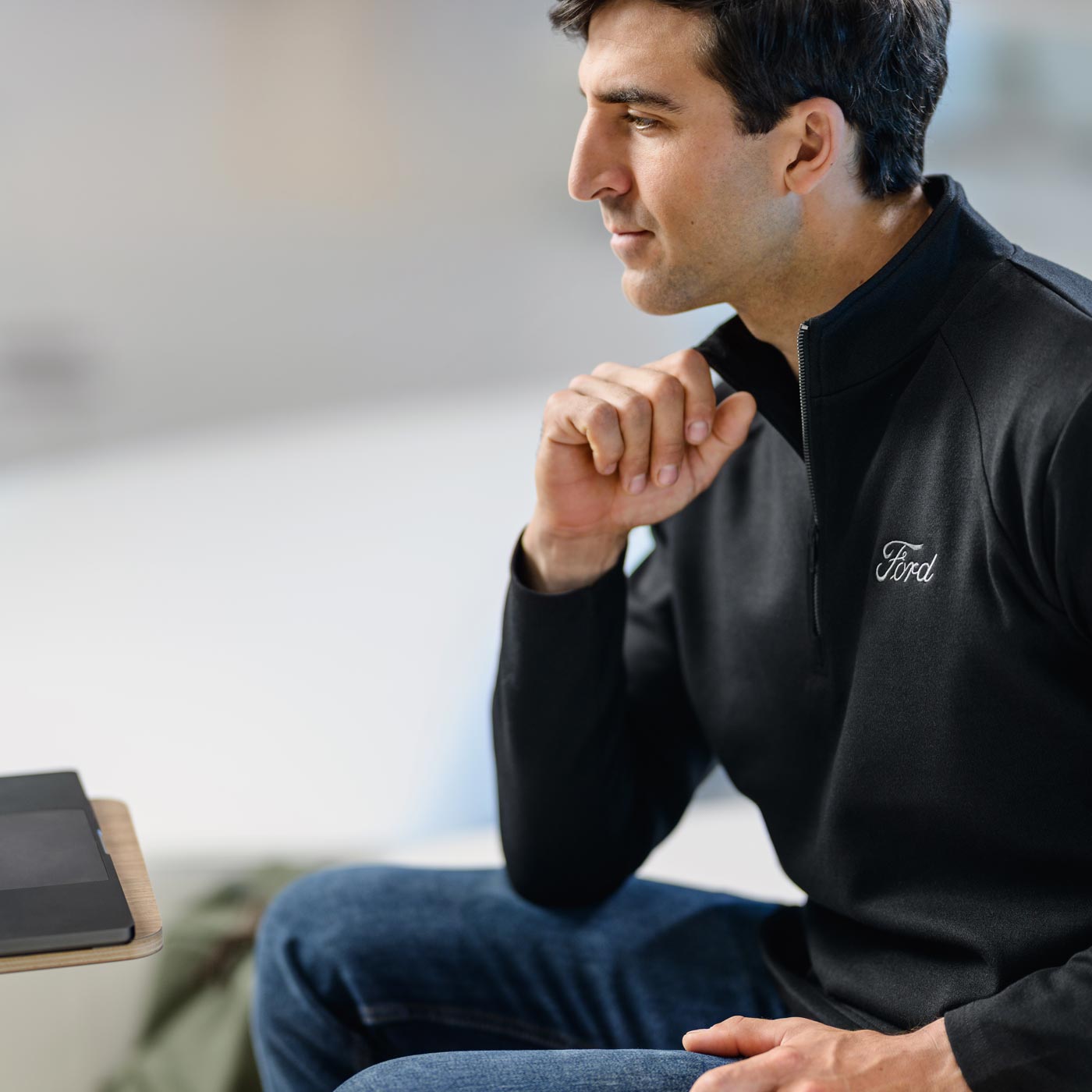 Man wearing a black jacket with a Ford logo, sitting indoors.