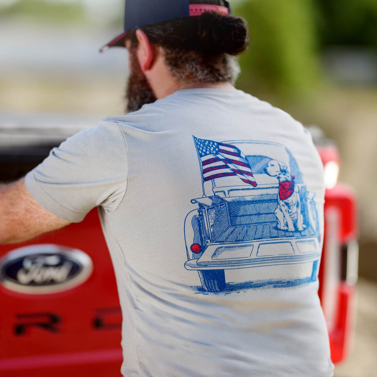 Person wearing a t-shirt with a graphic of a truck and American flag, standing next to a red Ford vehicle.