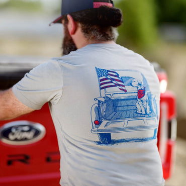 Person wearing a t-shirt with a graphic of a truck and American flag, standing next to a red Ford vehicle.