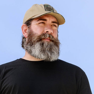 Man with a beard wearing a beige cap with built ford tough camo logo against a light blue background