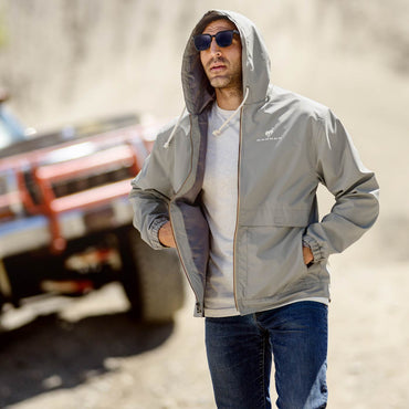 Man wearing a gray jacket with a hood, sunglasses, and a white shirt outdoors.