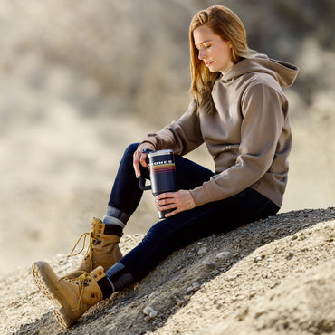 Person sitting outdoors holding a insulated tumbler, wearing a hoodie and boots.
