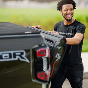Man opening the tailgate of a Ford Raptor truck with a blurred green field in the background