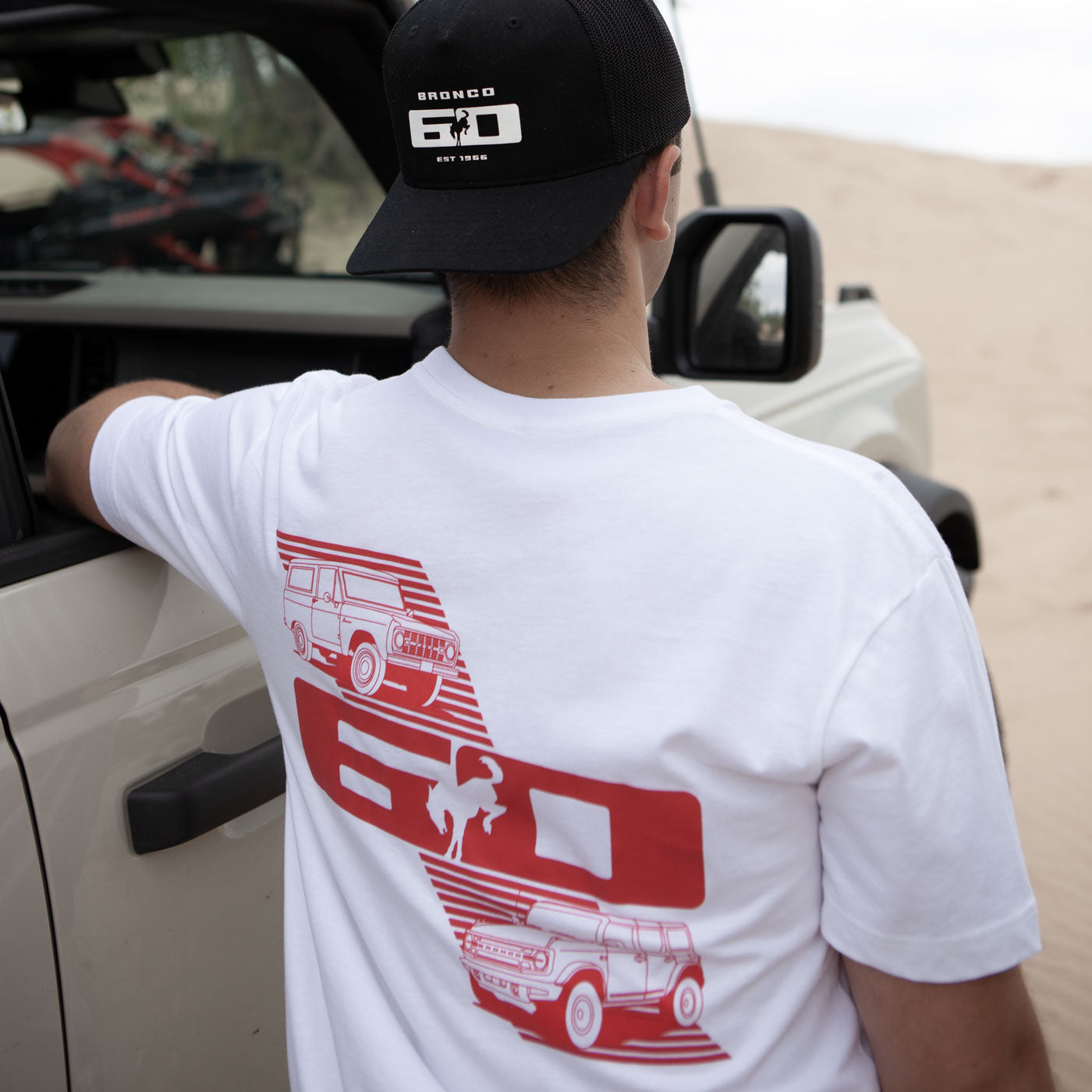 Person wearing a white t-shirt with red graphics and a black cap, standing next to a vehicle.
