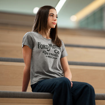 Woman wearing a gray t-shirt with 'Ford Motor Co Detroit Mich' text, sitting on steps.