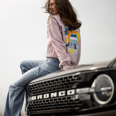Person wearing a pink hoodie with a bronco graphic design sitting on a car