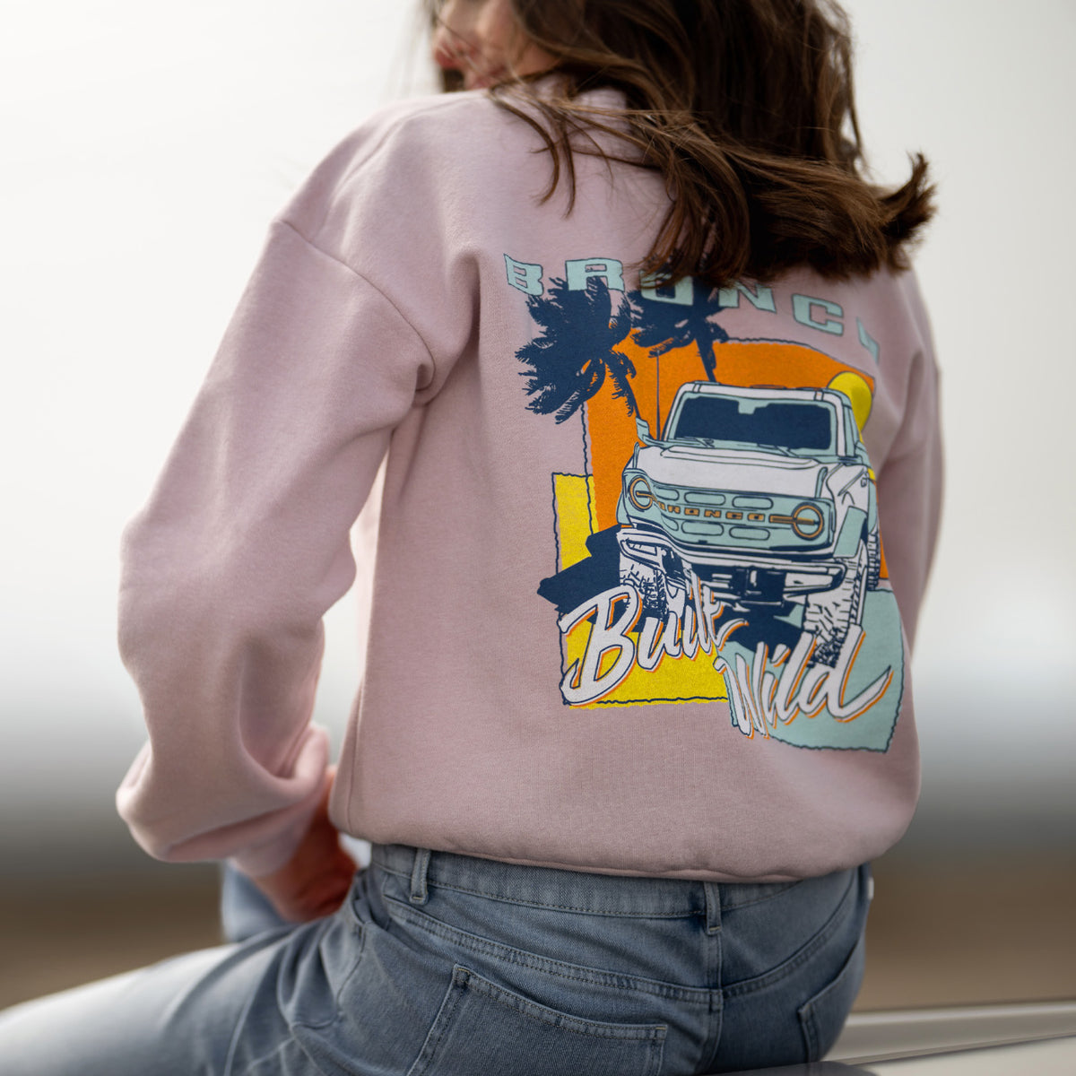 Person wearing a pink sweatshirt with a bronco graphic design on the back, sitting on a couch.