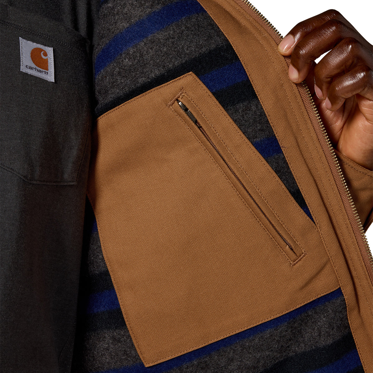 Close-up of a Carhartt jacket showing the interior pocket, brown waxed canvas flap, striped wool-blend lining in gray and navy, and a hand opening the zipper.