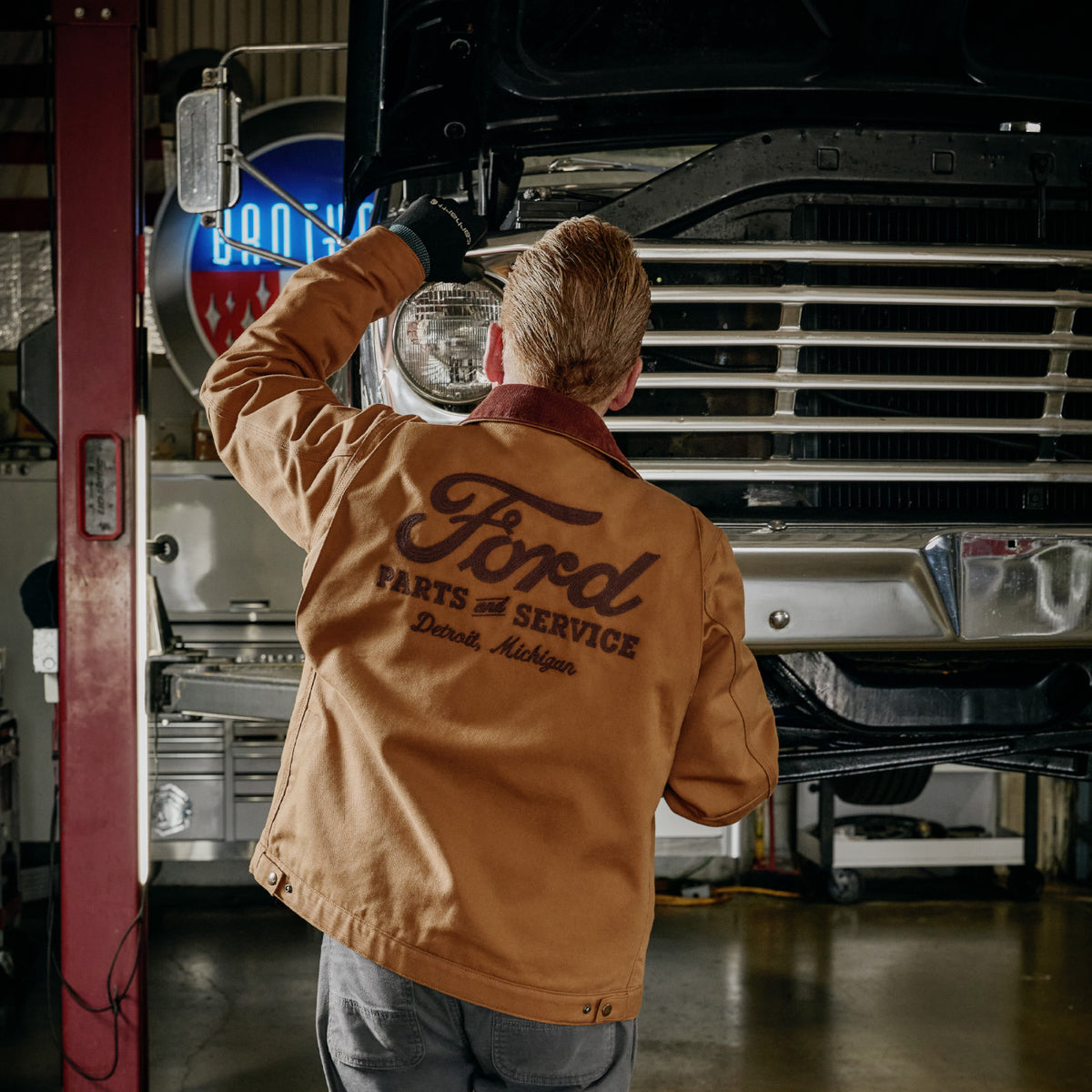 A man seen from behind wearing a tan Ford Parts & Service work jacket, standing in an auto repair garage under a raised vintage truck.