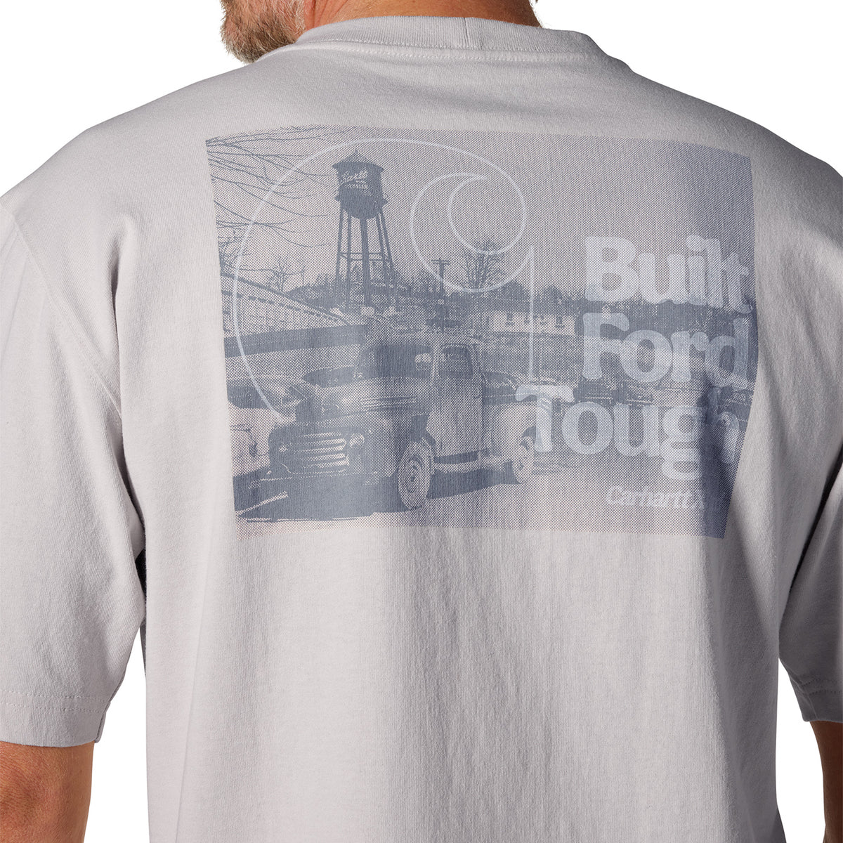 Close-up of the back of a light gray t-shirt worn by a model, featuring a faded blue-gray graphic print of a vintage Ford truck and water tower with the text 'Built Ford Tough' and 'Carhartt'.