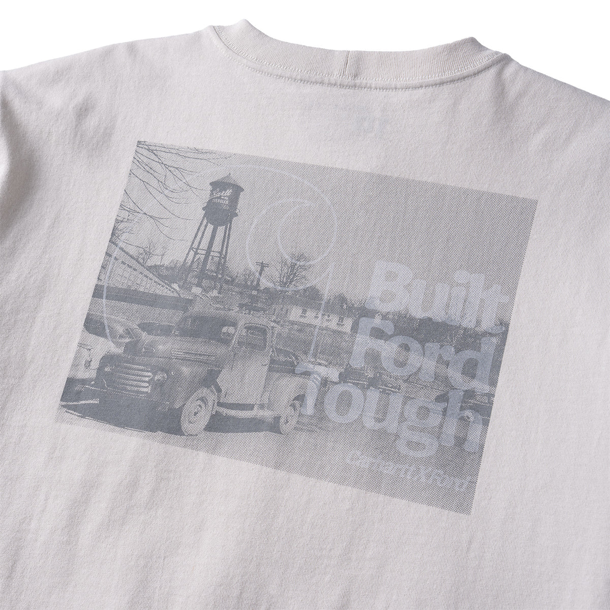 Close-up of the back of a light gray or off-white T-shirt with a large rectangular grayscale graphic print. The print shows an old pickup truck, a water tower, and faded text reading 'Built Ford Tough' with 'Carmen Rivera' below.