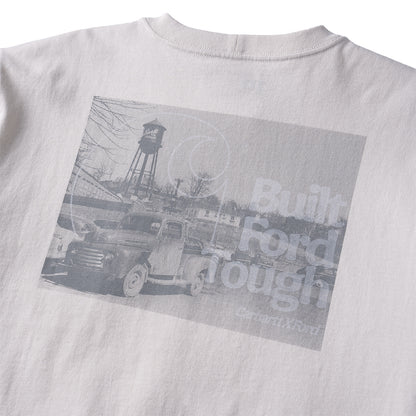 Close-up of the back of a light gray or off-white T-shirt with a large rectangular grayscale graphic print. The print shows an old pickup truck, a water tower, and faded text reading 'Built Ford Tough' with 'Carmen Rivera' below.