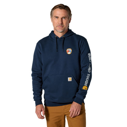A man wearing a navy blue Carhartt hoodie with a small chest logo and a large vertical logo print on the left sleeve, paired with brown pants, against a plain white background.