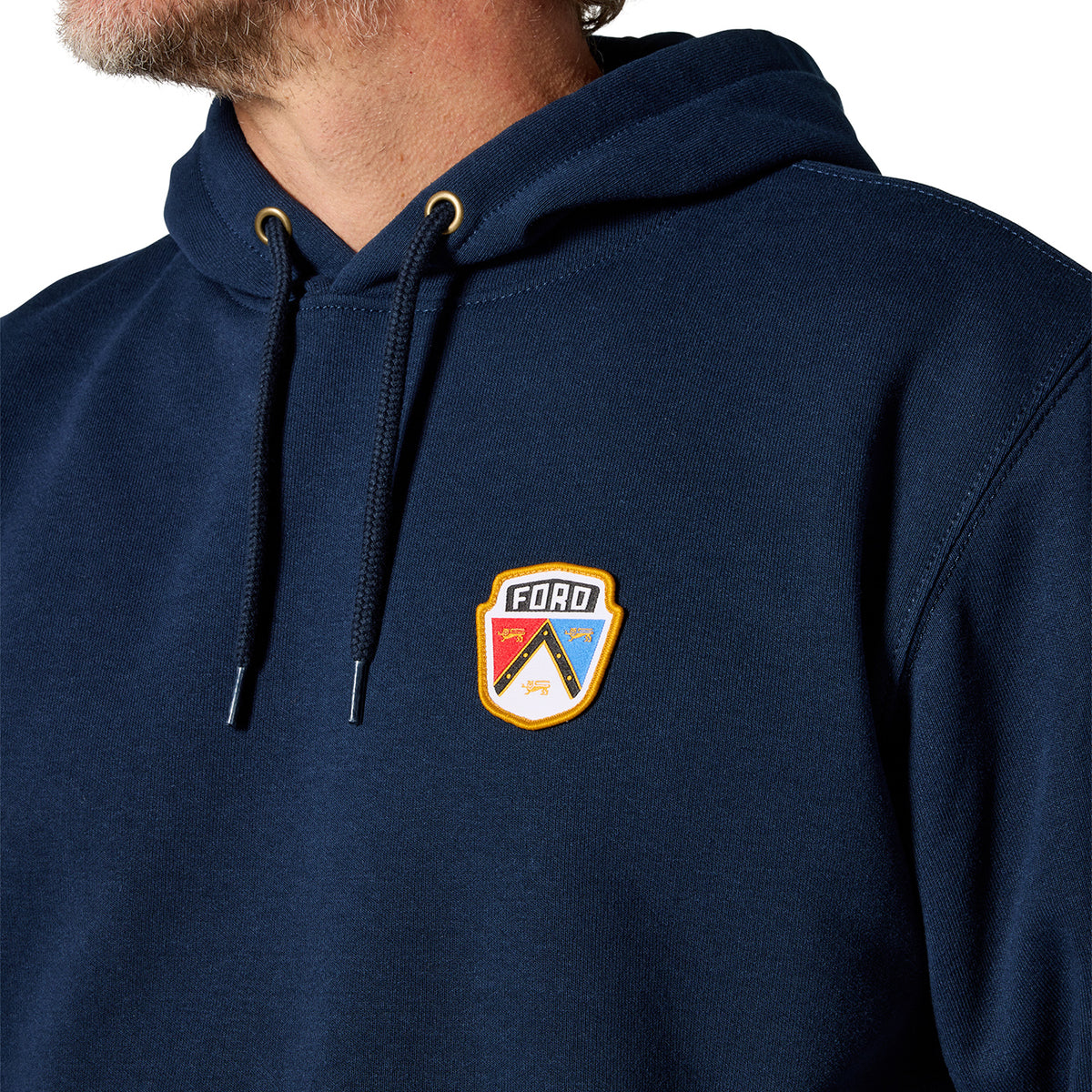 Close-up of a person wearing a navy pullover hoodie with a small shield-shaped FORD logo patch on the left chest. The hoodie has black drawstrings with gold eyelets and a white, red, blue, and yellow logo patch on the chest.