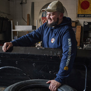 A smiling man in a workshop wearing a navy blue hoodie with 'BUILT FORD TOUGH' printed on the sleeve and a tan cap, leaning on a black metal case next to a tire.
