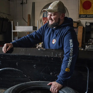 A smiling man in a workshop wearing a navy blue hoodie with 'BUILT FORD TOUGH' printed on the sleeve and a tan cap, leaning on a black metal case next to a tire.