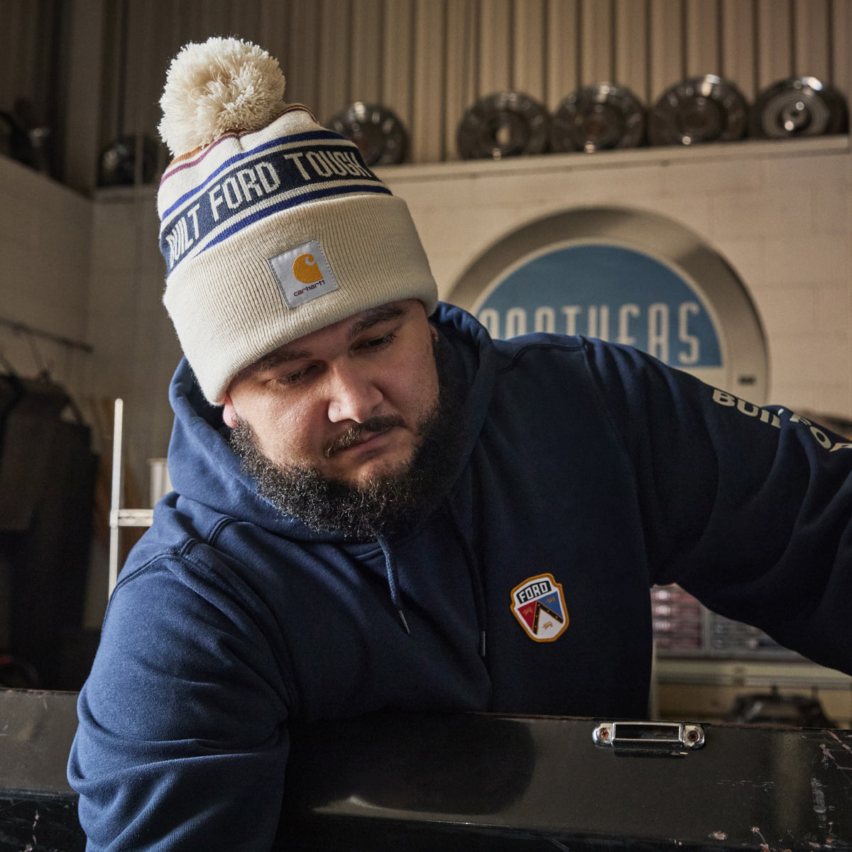 A man wearing a navy blue hoodie with a Carhartt logo patch and a beige beanie with a pom-pom and 'COLD FORD TOYS' text, leaning over a workbench in an automotive shop.