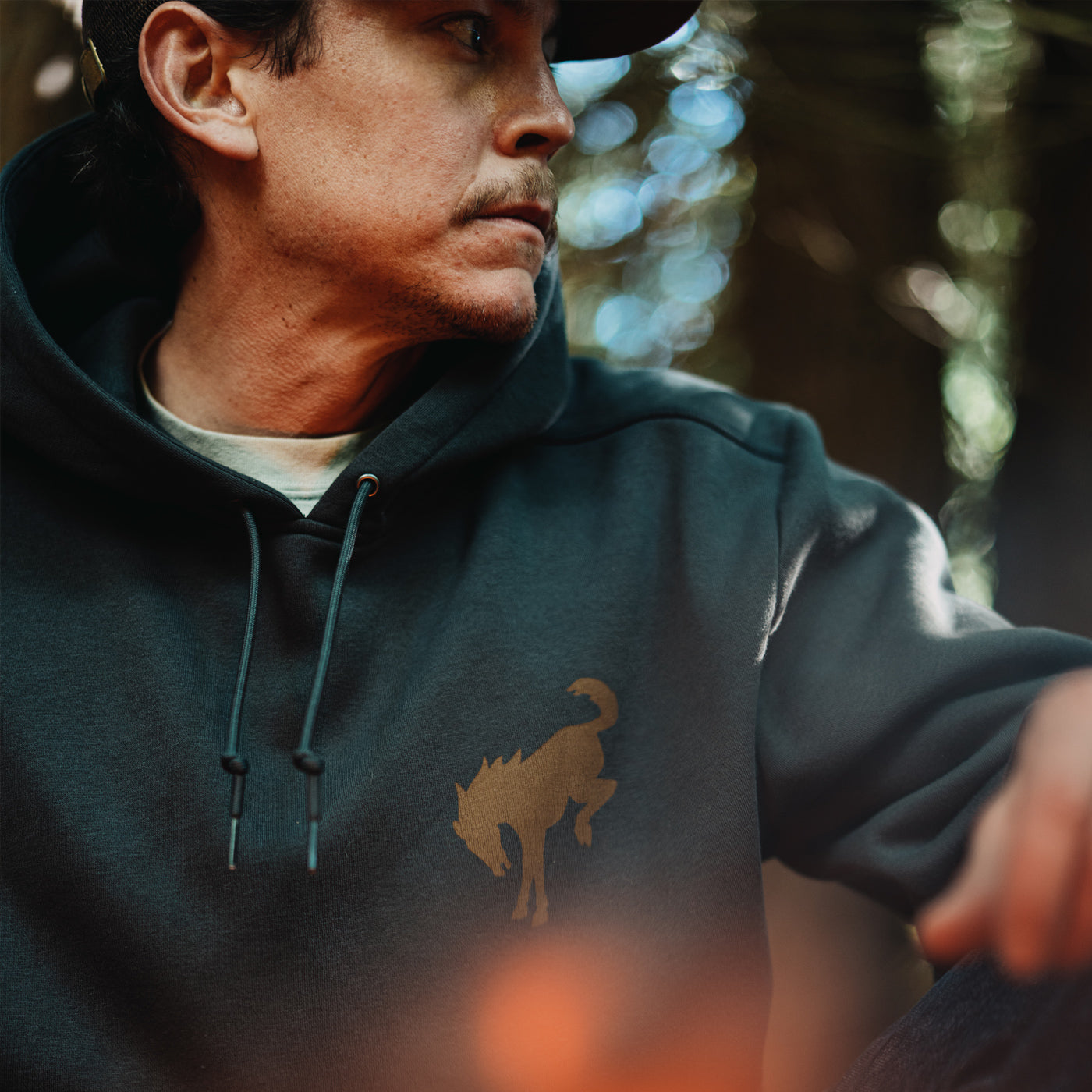 Man wearing a dark hoodie with a logo, standing outdoors with a blurred natural background