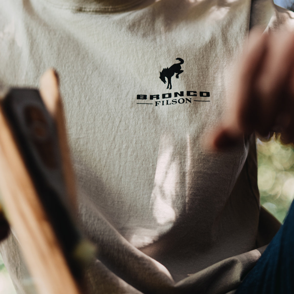 Person wearing a shirt with 'Bronco Filson' logo, holding a tool.