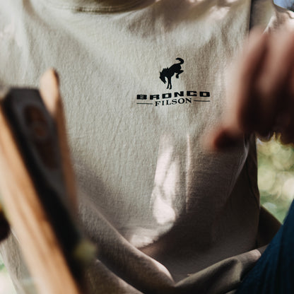 Person wearing a shirt with 'Bronco Filson' logo, holding a tool.