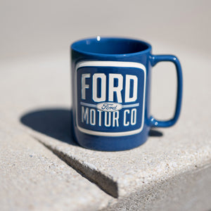 FRONT of a navy blue vintage text ford motor co ceramic mug sitting on a stone ledge