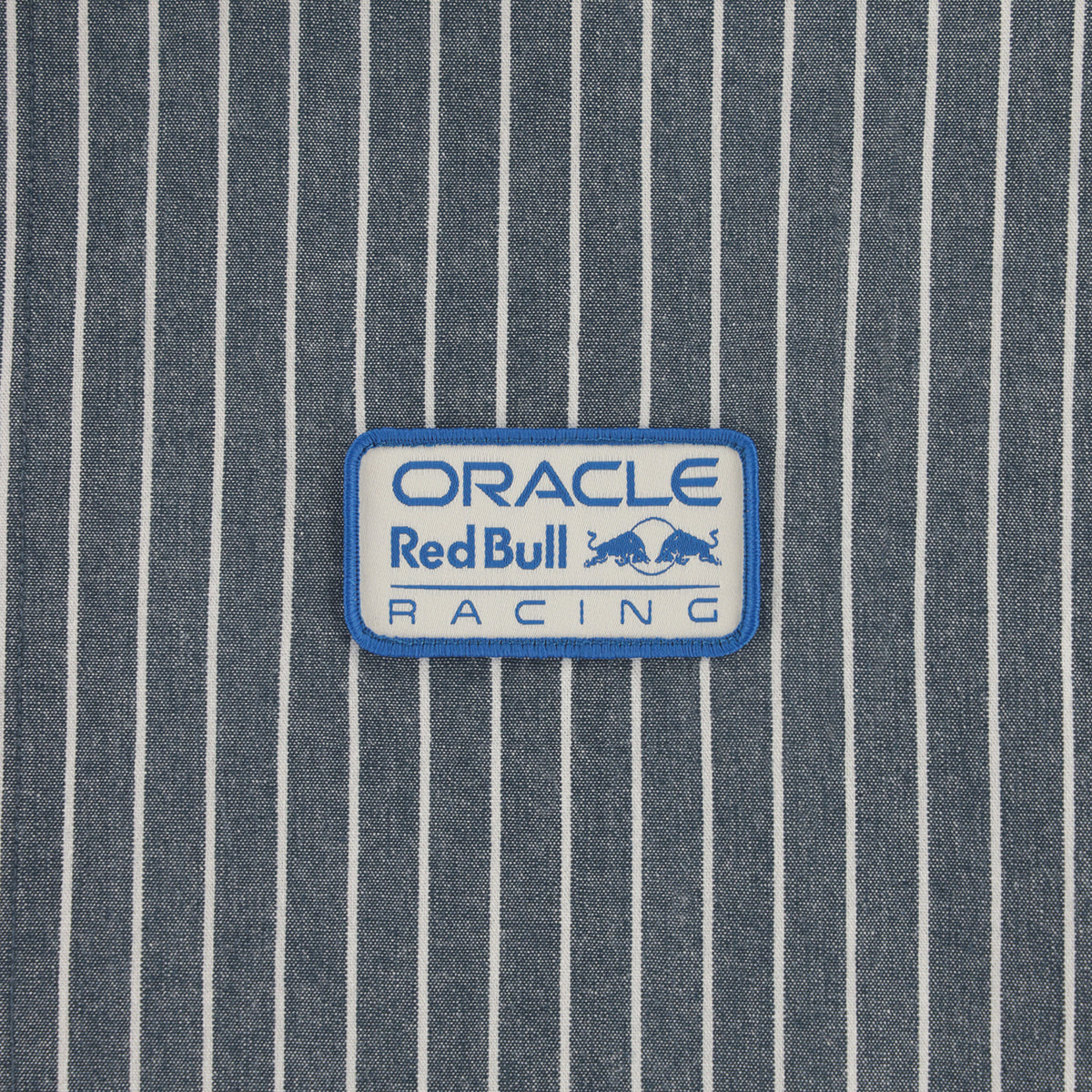 A rectangular embroidered patch with a white background and blue border, featuring the text 'ORACLE Red Bull RACING' and the Red Bull logo, placed on a blue and white vertically striped fabric background.