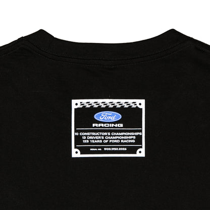 Close-up of the back of a black t-shirt featuring a white rectangular graphic patch with the blue Ford Racing logo and text celebrating 125 years of Ford Racing.