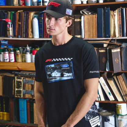 A man wearing a black Formula 1 t-shirt with a Ford race car graphic and a matching black cap, standing in front of a bookshelf filled with books and bottles.