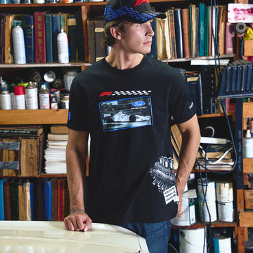 A man wearing a black Ford racing t-shirt and a blue patterned cap stands in front of a bookshelf filled with books and cans, leaning on a white car hood.