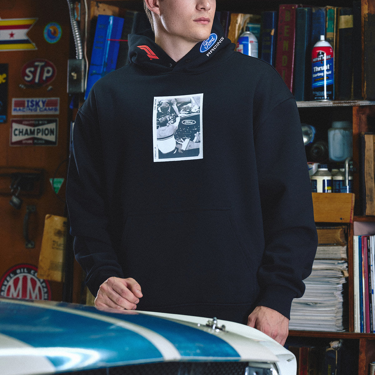 A man wearing a black pullover hoodie with a square black-and-white graphic print on the chest, standing in a garage-like setting with a blue and white car hood in the foreground.