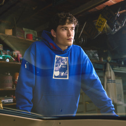 A young man wearing a bright blue hoodie with a graphic print on the chest, standing in a cluttered garage or workshop environment.