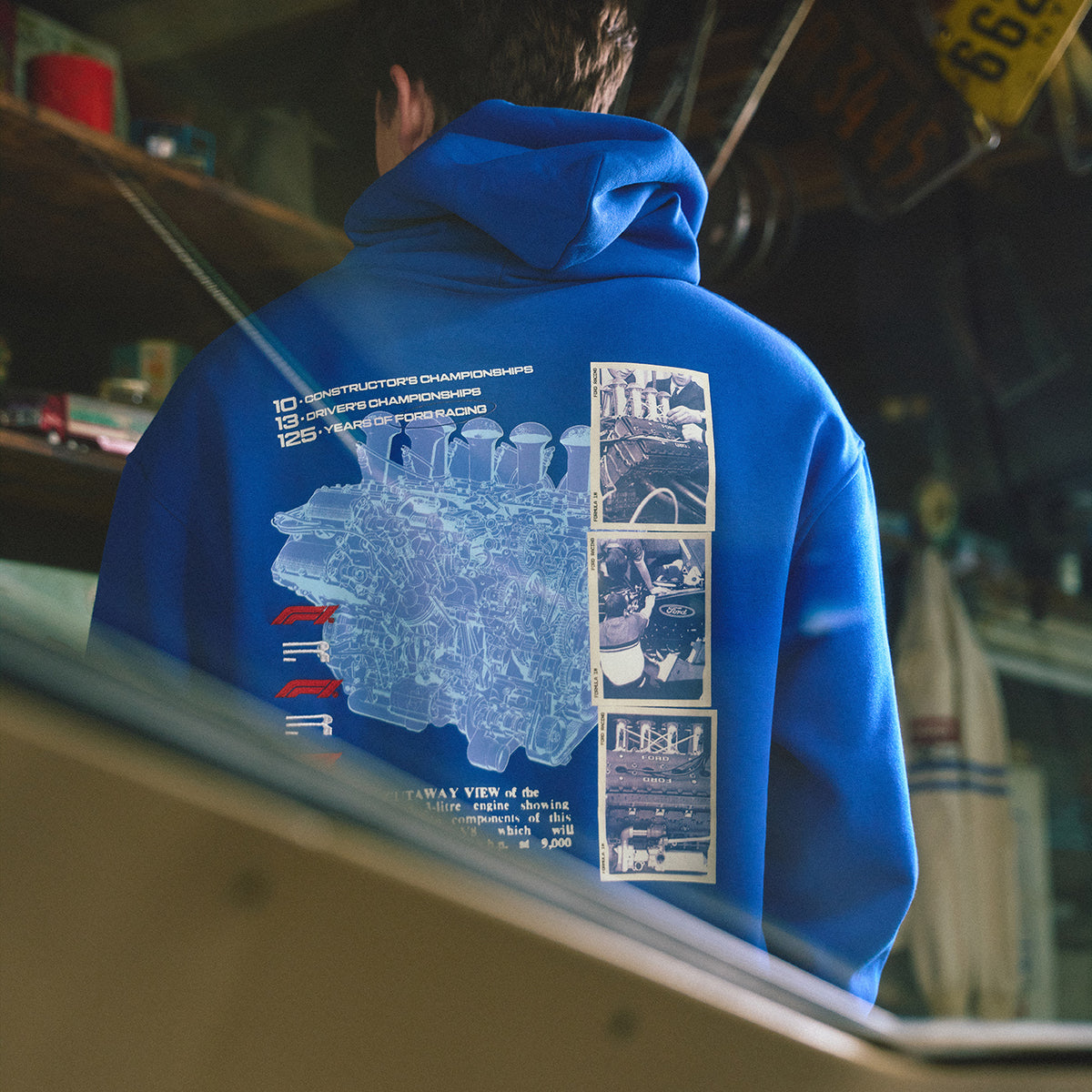 A person viewed from behind wearing a bright blue hoodie with a large technical-style back graphic in pale blue and white, featuring text and small inset photos. The setting appears to be an indoor shop or garage with shelves and signage in the background.