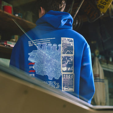 A person viewed from behind wearing a bright blue hoodie with a large technical-style back graphic in pale blue and white, featuring text and small inset photos. The setting appears to be an indoor shop or garage with shelves and signage in the background.